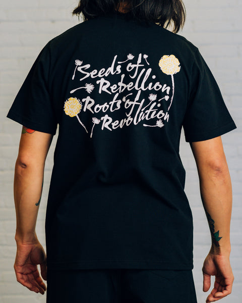 Back view of white tee with black script “Seeds of Rebellion Roots of Revolution” and yellow and black dandelions around it.