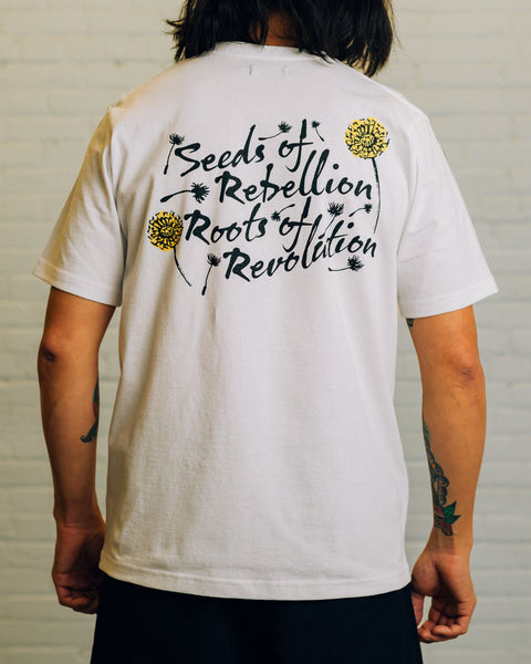 Back view of white tee with black script “Seeds of Rebellion Roots of Revolution” and yellow and black dandelions around it.