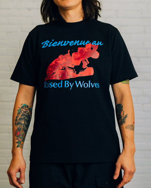 Front of white tee white green cursive text saying “Bienvenue au” and text saying “Raised By Wolves” and blue race cars.