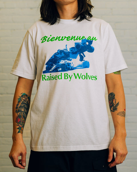 Front of white tee white green cursive text saying “Bienvenue au” and text saying “Raised By Wolves” and blue race cars.