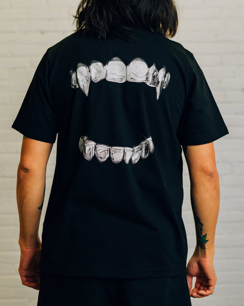 Back view of a white tee with silver grills screen printed on it. The front has “RAISED BY WOLVES” printed in black.