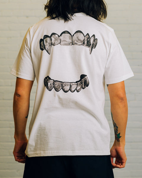 Back view of a white tee with silver grills screen printed on it. The front has “RAISED BY WOLVES” printed in black.