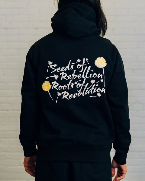 Back of an olive hoodie with script “Seeds of Rebellion Roots of Revolution” and yellow and white dandelions around it.