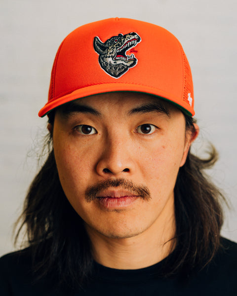 Front view of an orange trucker hat with embroidered wolf logo and mesh backing. The side view features ’47 logo in white.