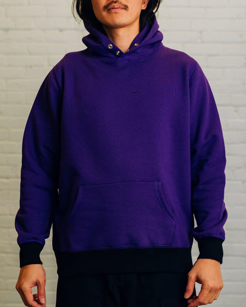 Front of a purple hoodie with two silver snap buttons at the neck. On the hood “Raised By Wolves” is embroidered in white.