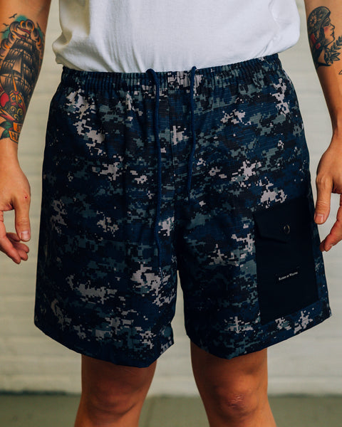 Front view of men’s shorts in a green digicam pattern with a black snap pocket near the bottom. Raised By Wolves is printed on the pocket in white.