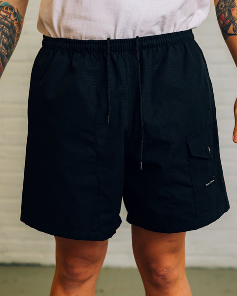 Front view of men’s shorts in a green digicam pattern with a black snap pocket near the bottom. Raised By Wolves is printed on the pocket in white.